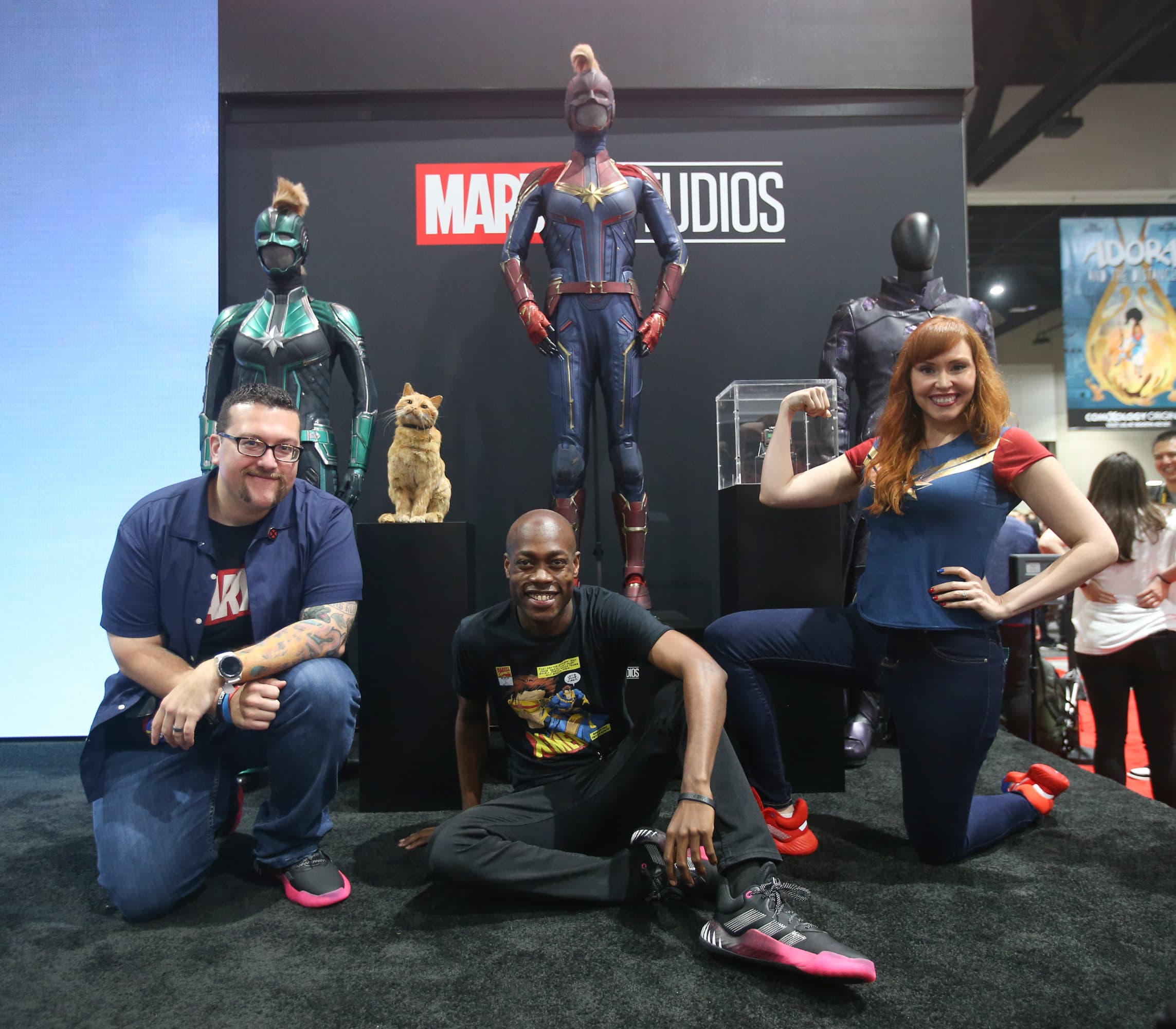 From left to right: Marvel Live Hosts Ryan "Agent M" Penagos, Langston Belton, and Lorraine Cink pose in front of costumes from Marvel Studios' Captain Marvel