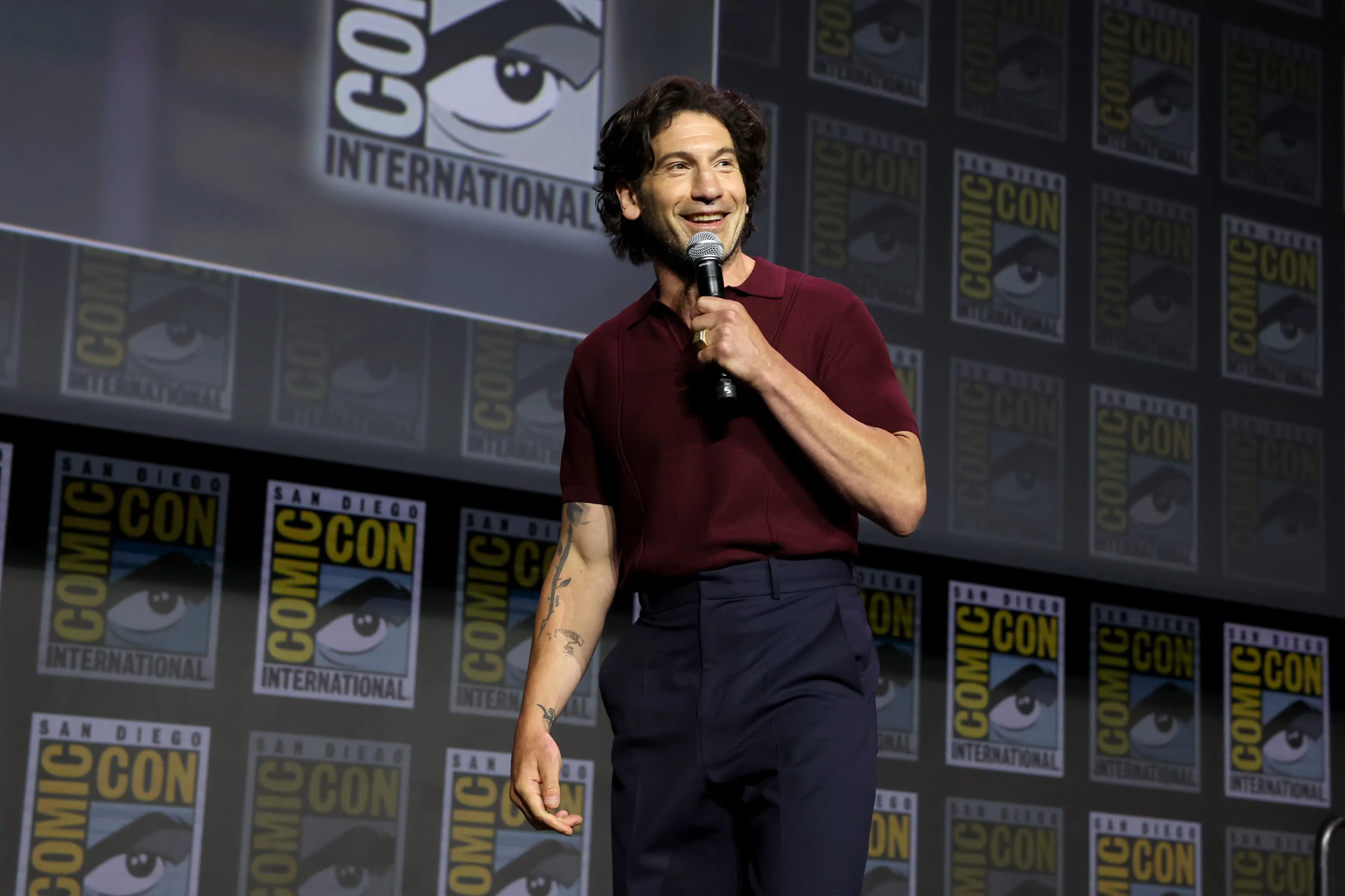 A medium full shot captures actor Jon Bernthal standing on a stage during a San Diego Comic-Con panel. He is smiling warmly while holding a microphone in his left hand, positioned near his chest. Bernthal has dark, wavy, medium-length hair and a light beard. He is wearing a short-sleeved, maroon knit polo shirt with a classic collar, paired with dark navy blue trousers. A black belt with a silver buckle is visible at his waist, and he has several tattoos on his right forearm. The background consists of a massive, dark grey wall covered in a repeating grid pattern of the official "San Diego Comic-Con International" logo, which features an stylized eye inside a speech bubble. The stage lighting is soft, illuminating him from the front and creating a professional, event-focused atmosphere.